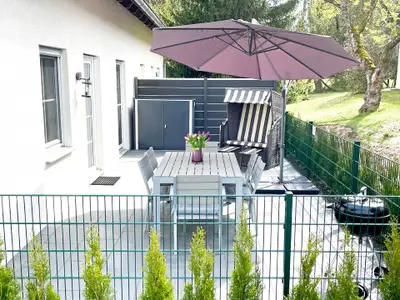 Umzäunte Terrasse mit  Grill, Strandkorb, 2 Sonnenliegen, Esstisch und 6 Stühlen.