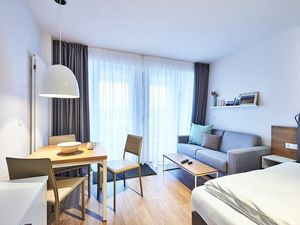 Wohn-Ess-Schlafbereich mit Sofa, Esstisch, Sitzgelegenheit und Doppelbett sowie Zugang zum Balkon