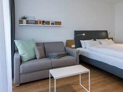 Wohn-Ess-Schlafbereich mit Sofa, Esstisch, Sitzgelegenheit und Doppelbett sowie Zugang zum Balkon