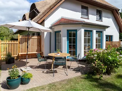 Reetgedecktes Haus mit Terrasse, Sonnenschirm, Sitzgruppe und blühenden Büschen.