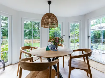 Essbereich mit rundem Holztisch und vier Stühlen, Blumenstrauß in der Mitte, große Fenster mit Blick in den Garten.