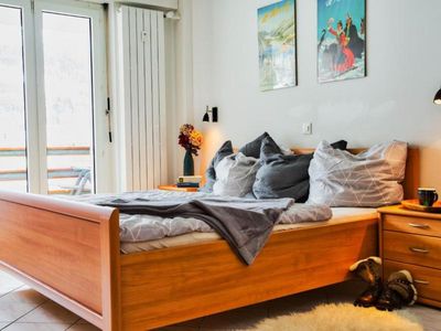 Schlafzimmer mit Bett, Schrank, TV und großer Fensterfront mit Zugang zum überdachten Balkon mit Blick auf St. Moritz.