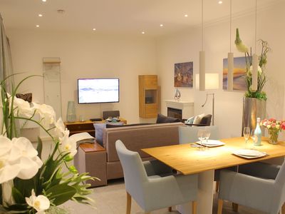 Wohnbereich mit Sofa, Sessel, Sideboard und TV, Esstisch mit Bestuhlung, Elektrokamin und offener Küche
