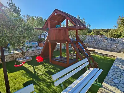 Die Spielzone für Kinder verfügt über ein Klettergerüst und Schaukeln auf grünem Untergrund, umgeben von Olivenbäumen.