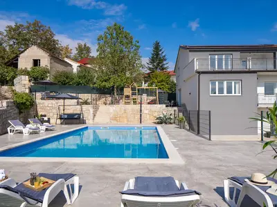 Genießen Sie den privaten 50 m2 großen Poolbereich mit bequemen Sonnenliegen vor der Kulisse des modernen Haupthauses und des erhöhten, von Steinmauern umgebenen Spielplatzes.